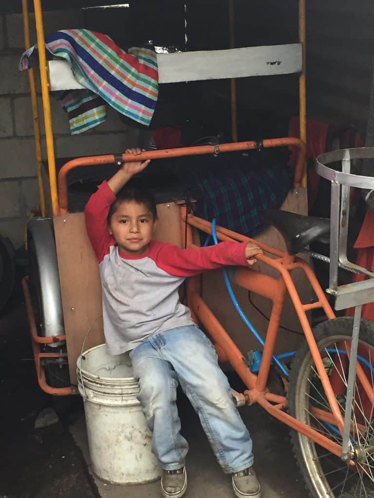 Manuel leans against the bicycle cart his mother uses to sell hot food. He rides on his mother’s back when she goes into the city.